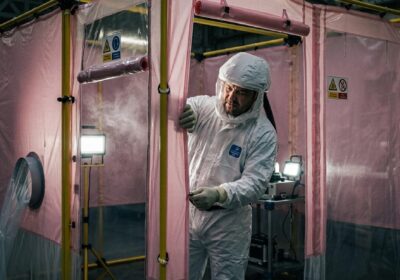 Technicien en combinaison de protection blanche ouvrant une tente rose isolée, équipements et projecteurs à l'intérieur.