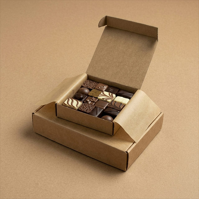 Boîte ouverte en carton contenant assortiment de chocolats variés décorés, posée sur une autre boîte en carton brun.