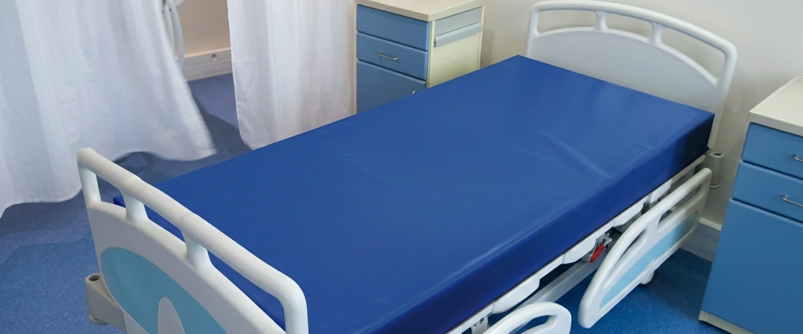 Lit d'hôpital vide avec matelas bleu, barrières blanches et chevets bleus, rideau d'intimité tiré en arrière.