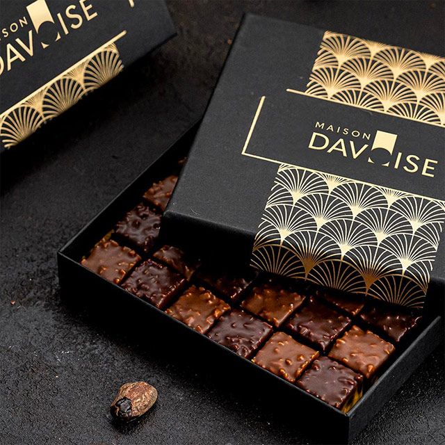 Boîte noire élégante ouverte de chocolats carrés glacés, avec couvercle décoré or à motifs éventails et une fève de cacao posée à côté.