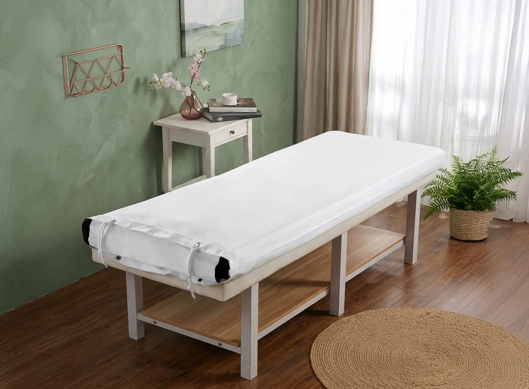 Table de massage avec housse blanche tendue, petite table d'appoint avec vase et livre, plante en pot et tapis rond sur sol en bois.