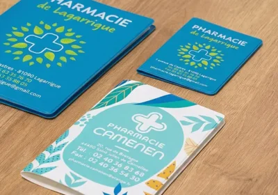 Trois carnets et cartes de visite bleu-vert pour des pharmacies, avec logo croix et motifs végétaux posés sur une table en bois.
