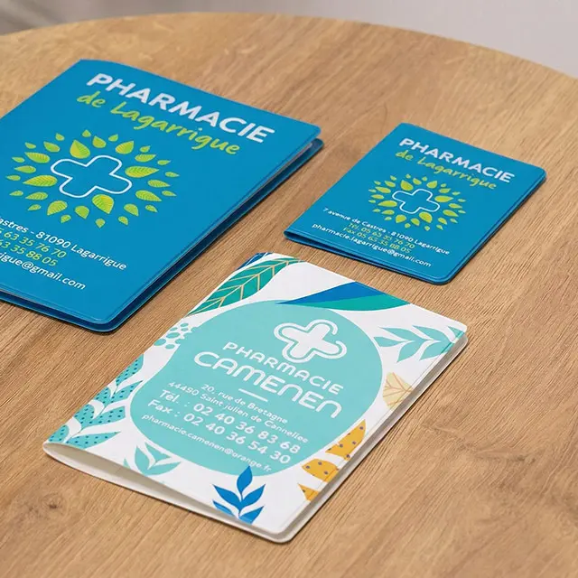 Trois carnets et cartes de visite bleu-vert pour des pharmacies, avec logo croix et motifs végétaux posés sur une table en bois.