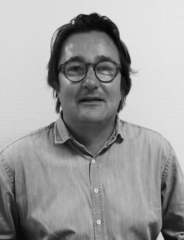 Homme portant des lunettes rondes et une chemise en denim, posé devant un mur texturé, regardant l'appareil.