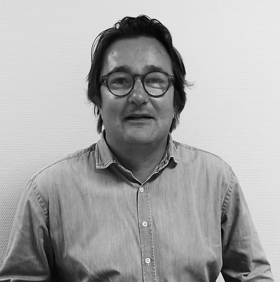 Homme portant des lunettes rondes et une chemise en denim, posé devant un mur texturé, regardant l'appareil.