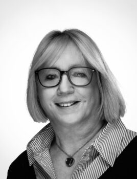Femme d’âge mûr souriante portant des lunettes carrées, coupe au carré blond, chemise rayée et pendentif cœur, fond neutre.