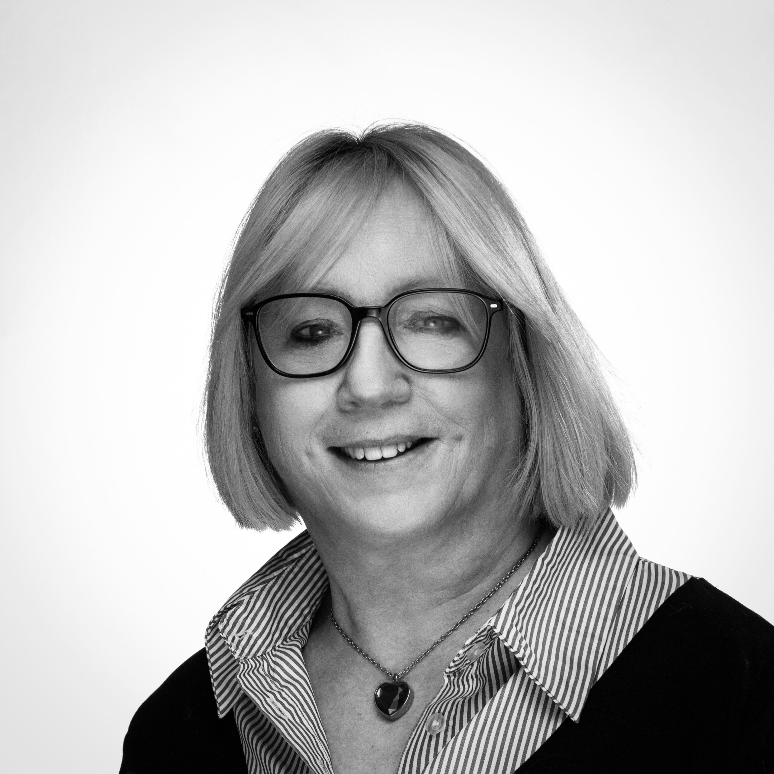 Femme d’âge mûr souriante portant des lunettes carrées, coupe au carré blond, chemise rayée et pendentif cœur, fond neutre.