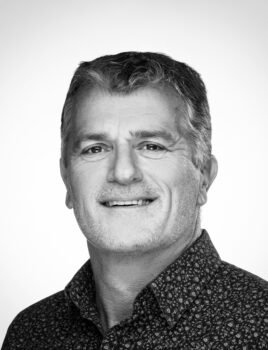 Homme d'âge moyen souriant en portrait noir et blanc, cheveux courts grisonnants et chemise à motifs.