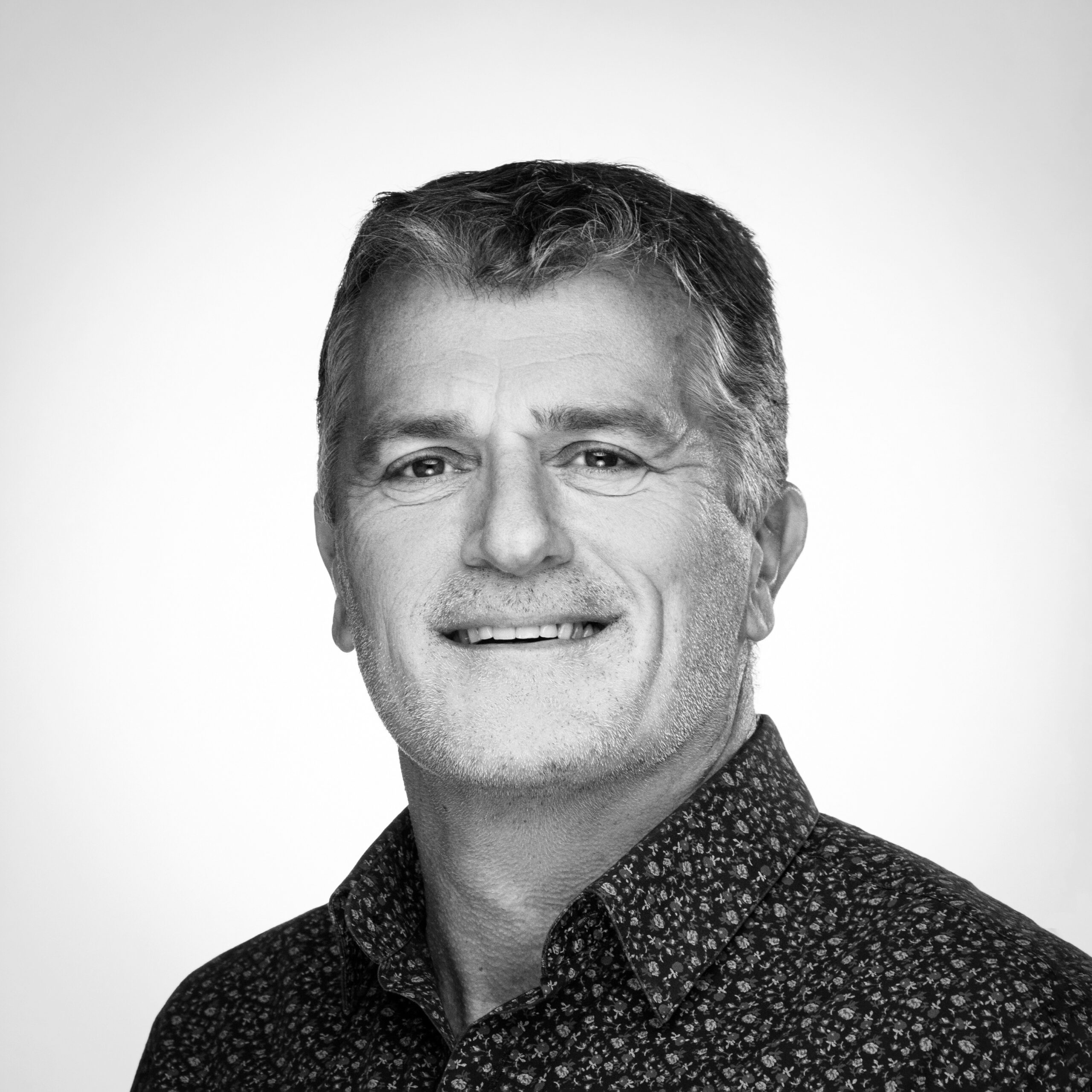 Homme d'âge moyen souriant en portrait noir et blanc, cheveux courts grisonnants et chemise à motifs.