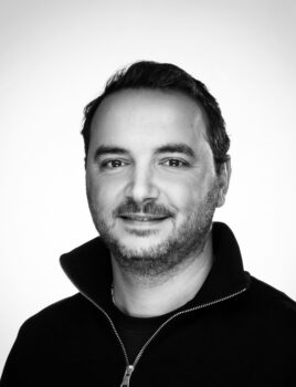 Portrait en noir et blanc d'un homme souriant au col montant zippé, regardant l'objectif, fond clair uniforme.
