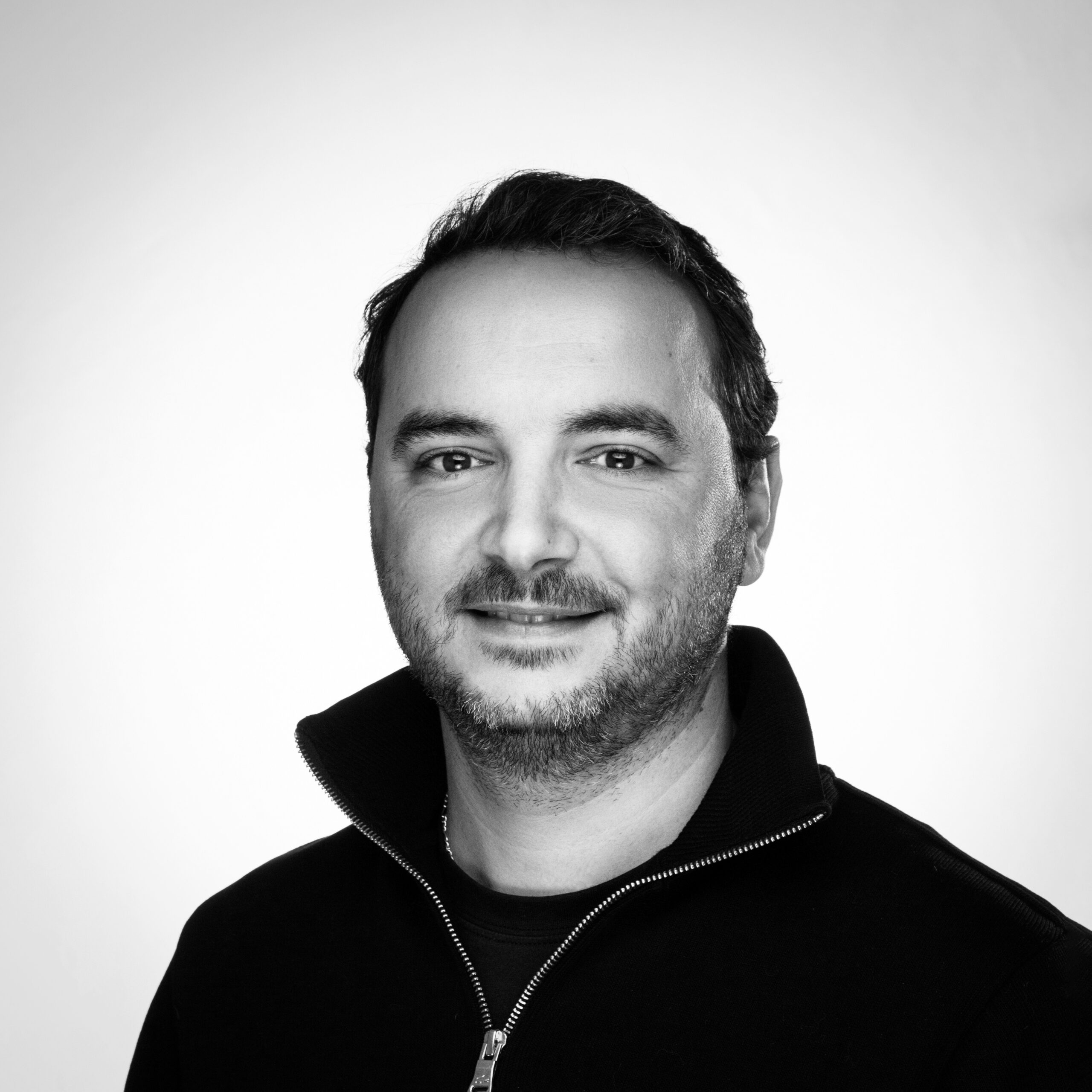 Portrait en noir et blanc d'un homme souriant au col montant zippé, regardant l'objectif, fond clair uniforme.