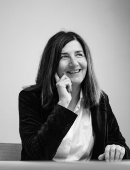 Femme souriante aux cheveux mi-longs, assise à une table, main posée sur le menton, vêtue d'une chemise blanche et d'une veste sombre.