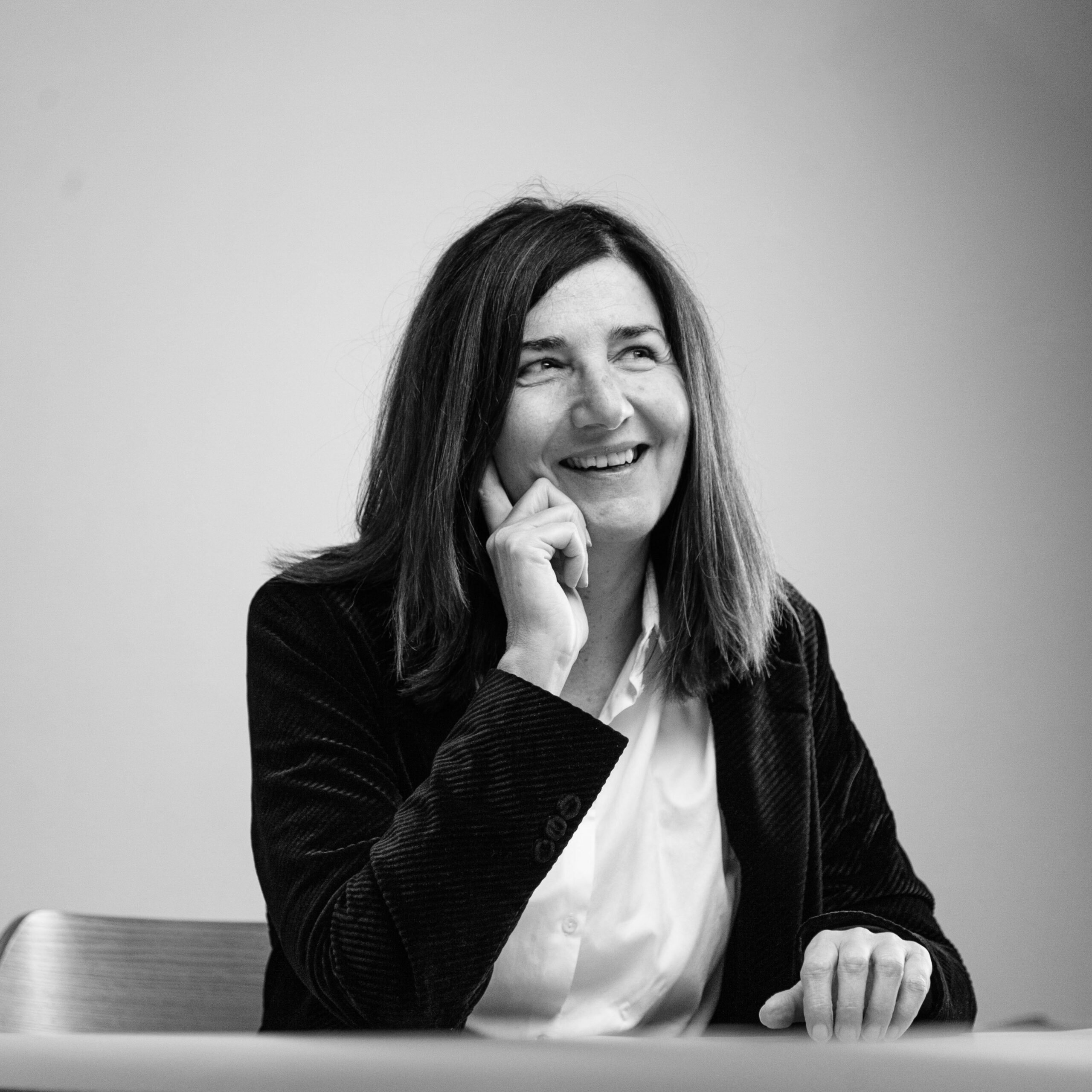 Femme souriante aux cheveux mi-longs, assise à une table, main posée sur le menton, vêtue d'une chemise blanche et d'une veste sombre.