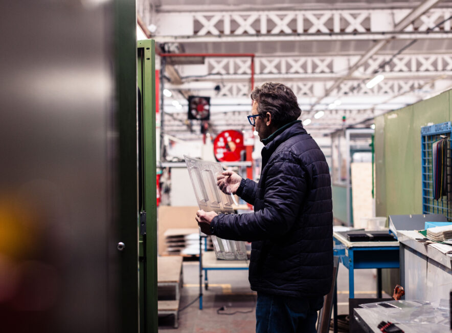 Ouvrier en veste matelassée examinant une pièce plastique transparente dans un atelier industriel.