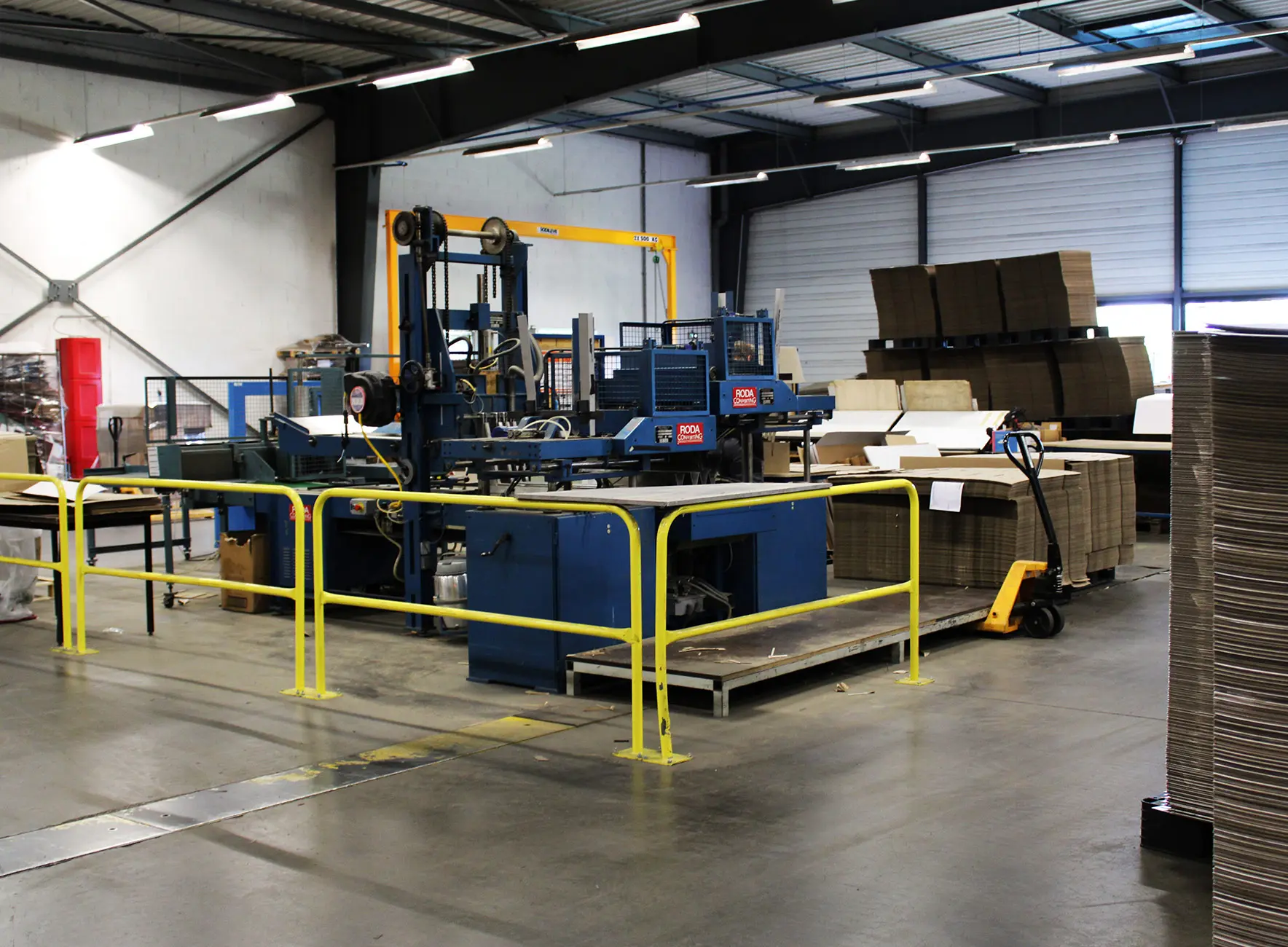 Atelier industriel avec machine d'emballage bleue entourée de barrières jaunes et piles de cartons sur palettes.