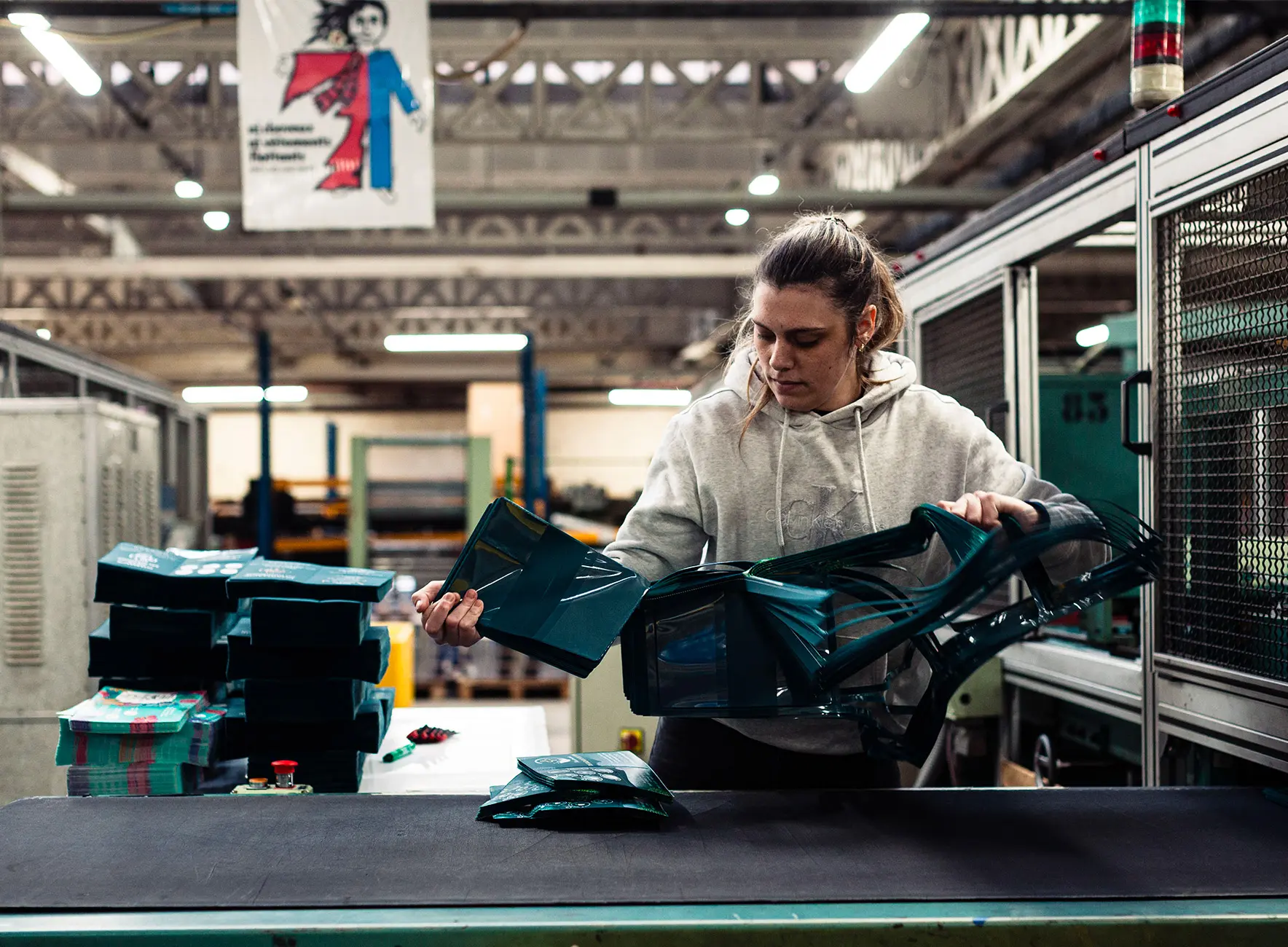 Une ouvrière dans un atelier pliant et empilant des pochettes plastiques vertes sur une table de travail industrielle.