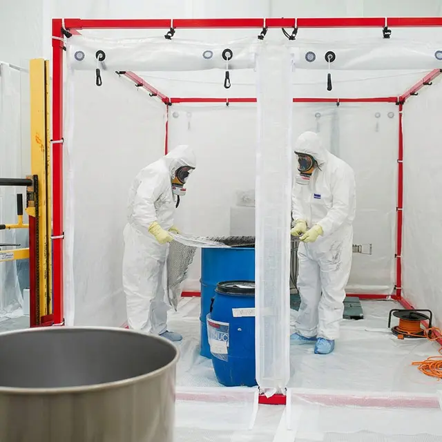 Deux techniciens en combinaisons blanches et masques respiratoires manipulant un fût bleu à l'intérieur d'une enceinte de confinement plastique.