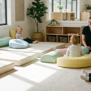 Enfant assis sur coussins colorés lisant un livre, autres tout-petits et une éducatrice souriante dans une salle de jeu lumineuse.