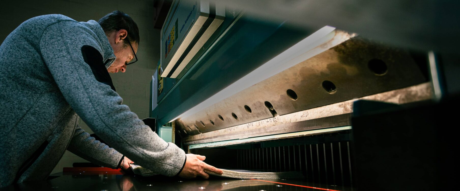 Homme penché réglant une grande guillotine industrielle, tenant une pile de feuilles près de la lame métallique.
