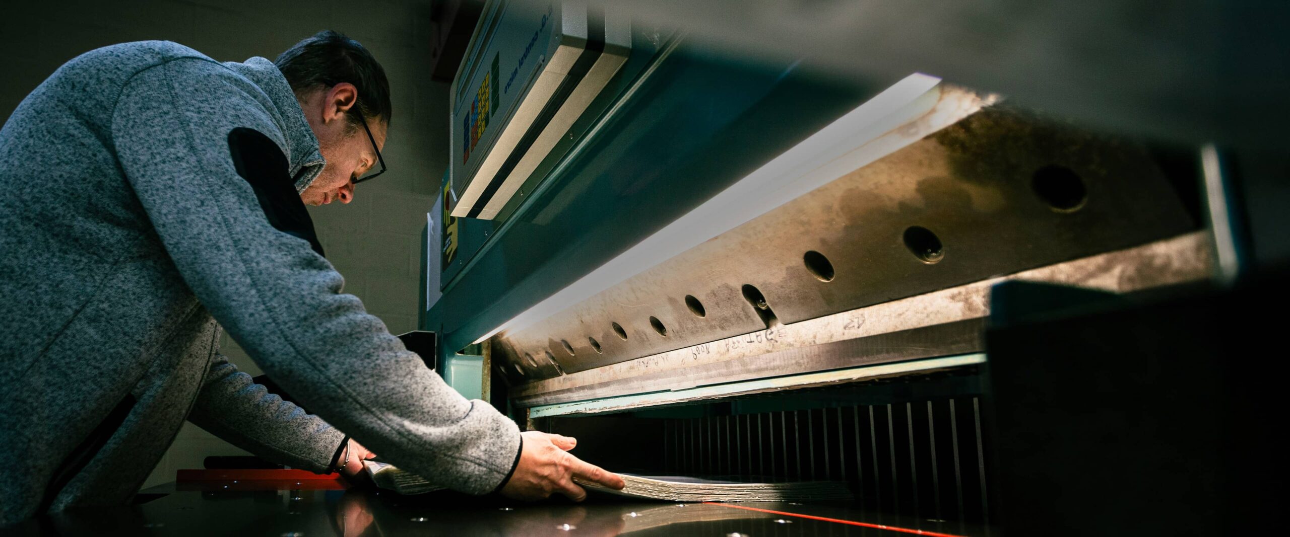 Homme penché réglant une grande guillotine industrielle, tenant une pile de feuilles près de la lame métallique.