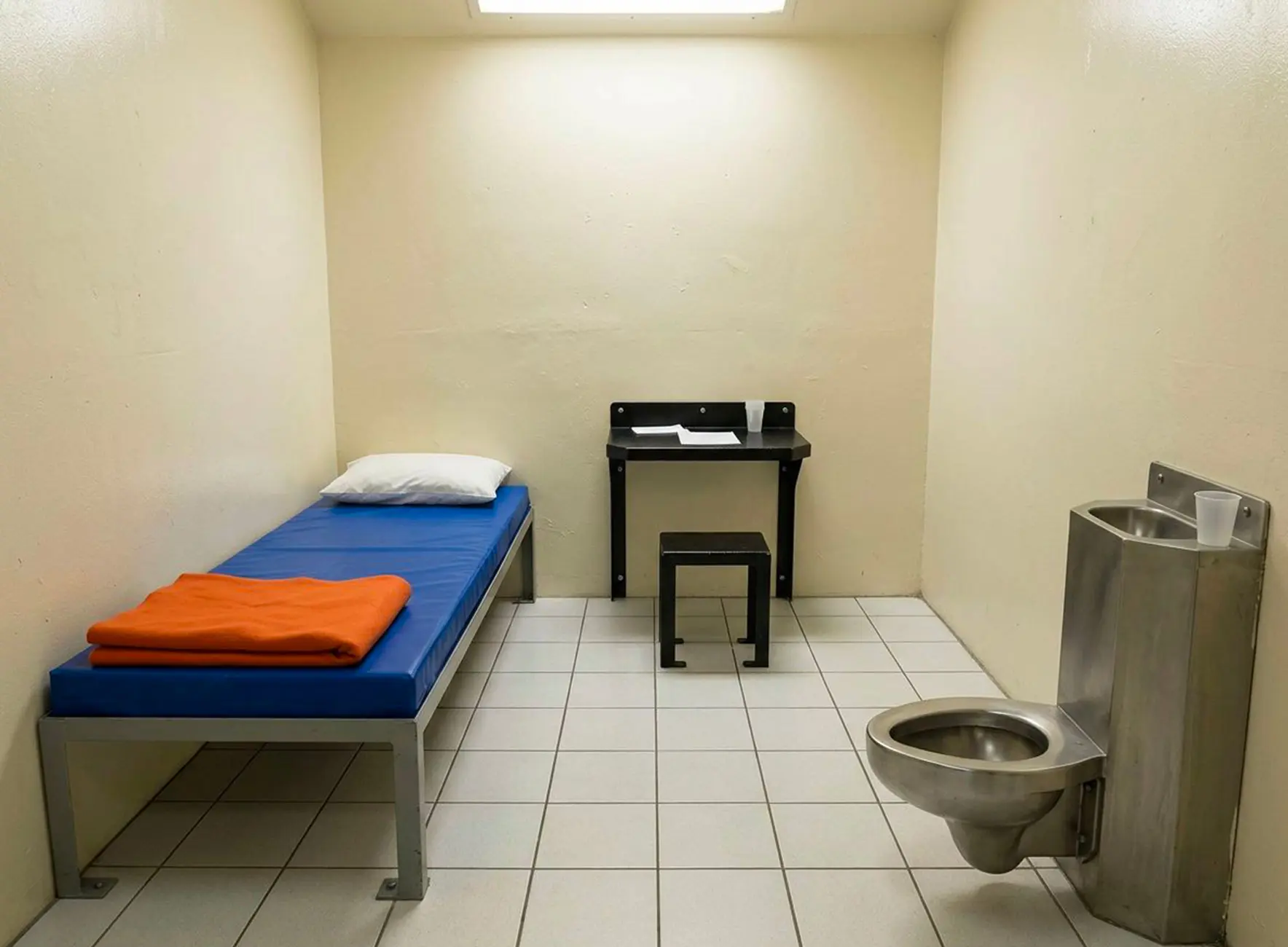 Cellule sobre avec lit métallique et matelas bleu, couverture orange, petite table et tabouret noirs, et toilette/lavabo en inox.