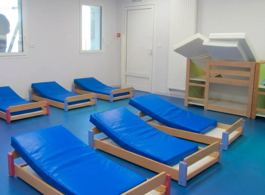 Petits lits inclinés en bois avec matelas bleus disposés sur un sol vinyle bleu dans une chambre d'enfants.