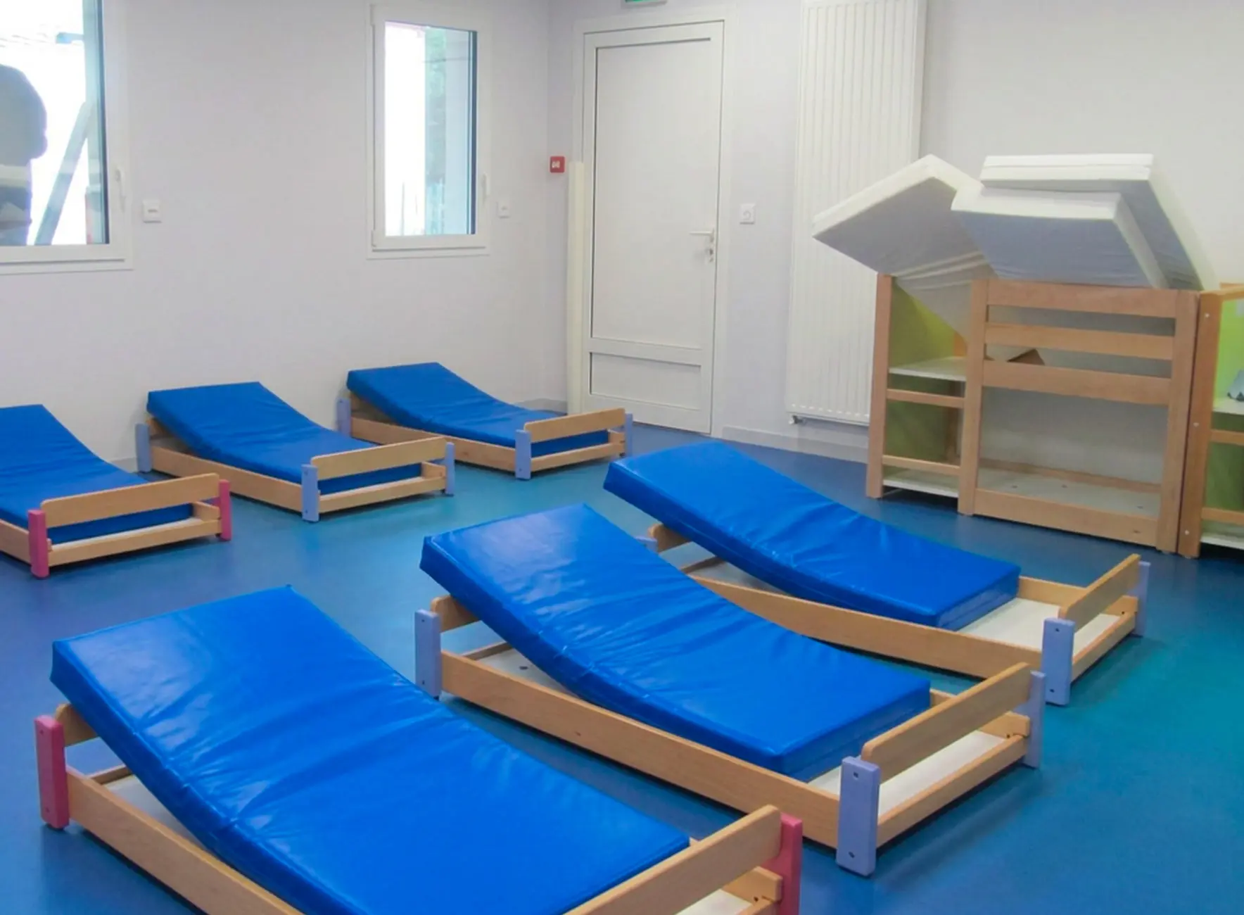 Petits lits inclinés en bois avec matelas bleus disposés sur un sol vinyle bleu dans une chambre d'enfants.