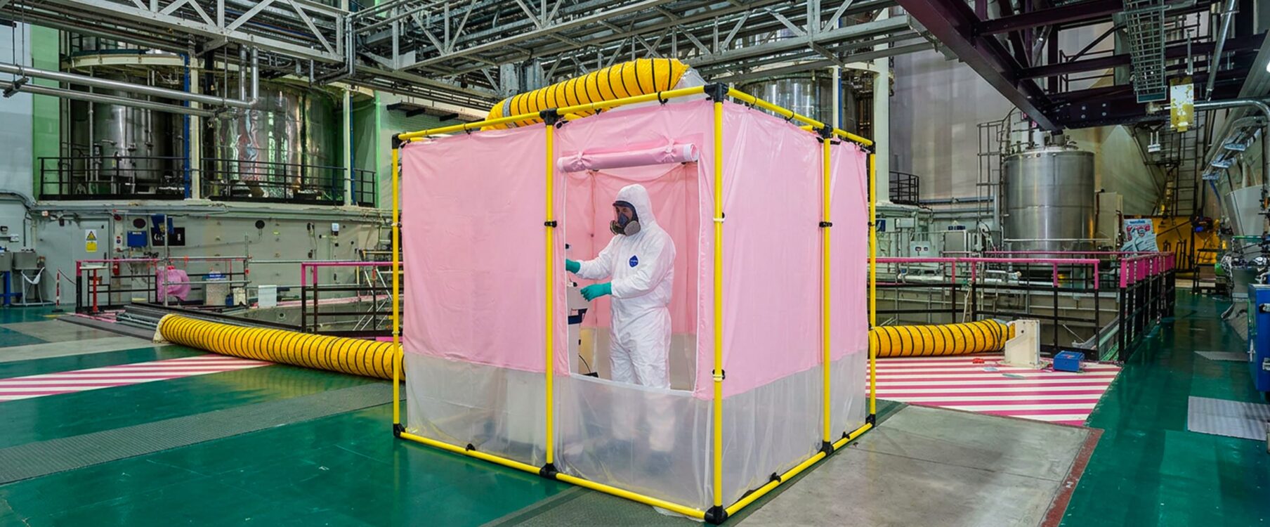 Technicien en combinaison intégrale et masque travaillant dans une cabine rose portable dans une grande enceinte industrielle avec tuyaux jaunes d'aération.