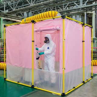 Technicien en combinaison intégrale et masque travaillant dans une cabine rose portable dans une grande enceinte industrielle avec tuyaux jaunes d'aération.