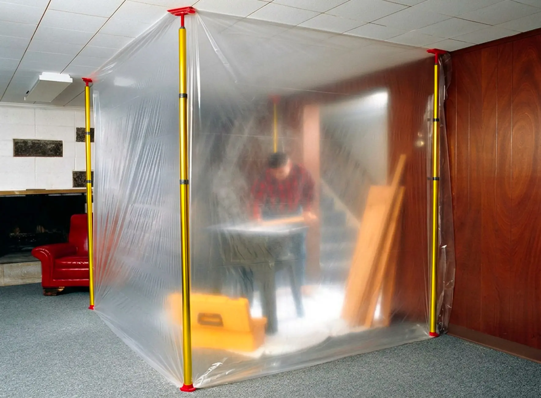 Cabine de chantier improvisée en plastique transparent maintenue par quatre tiges jaunes et pièces rouges, personne floue travaillant à l'intérieur.