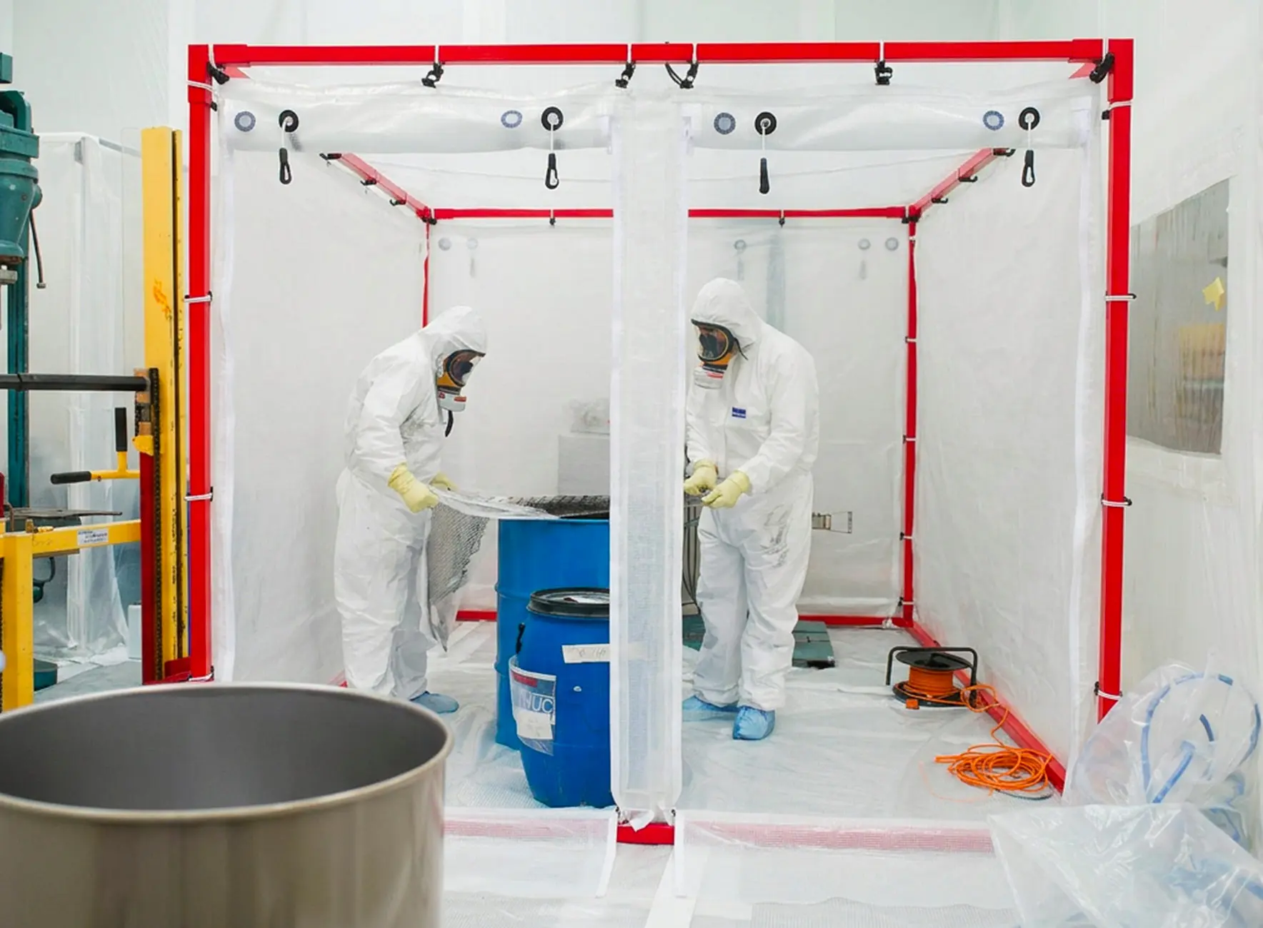 Deux techniciens en combinaisons blanches et masques respiratoires manipulant un fût bleu à l'intérieur d'une enceinte plastique protégée.