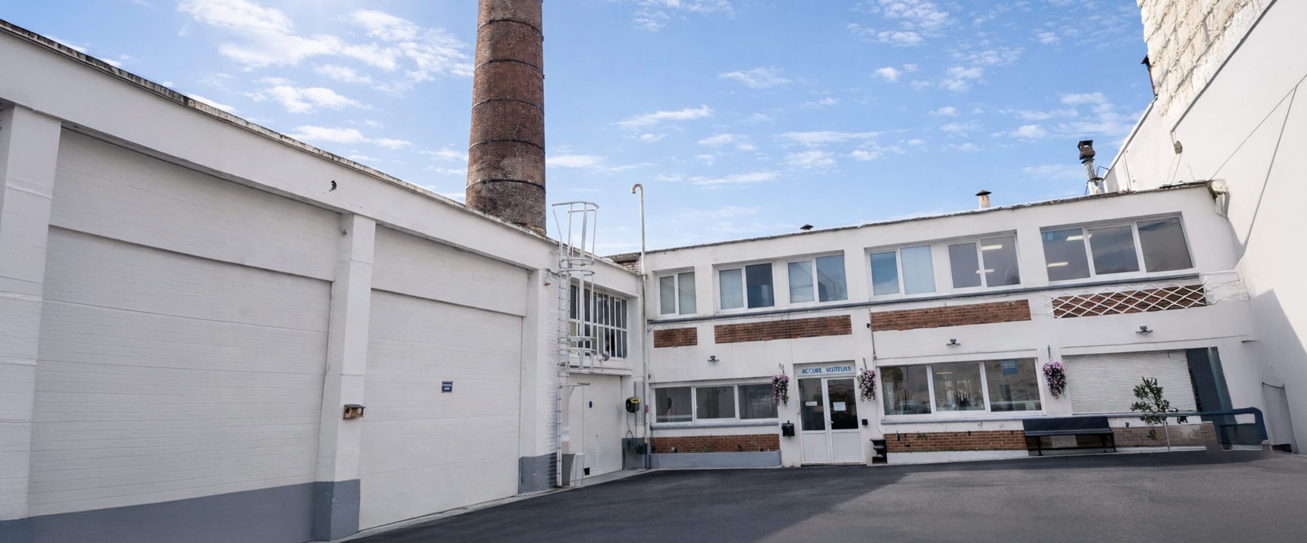 Cour intérieure d'un bâtiment industriel blanc à deux étages avec fenêtre, porte d'entrée, banc et une haute cheminée en brique en arrière-plan.