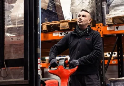 Ouvrier d'entrepôt portant gants et veste noire, tenant un transpalette rouge devant des étagères chargées de palettes emballées.
