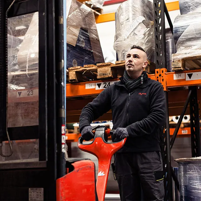 Ouvrier d'entrepôt portant gants et veste noire, tenant un transpalette rouge devant des étagères chargées de palettes emballées.