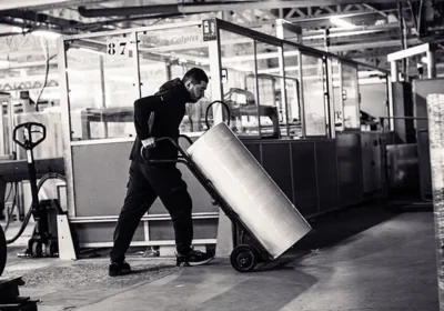 Employé poussant un transpalette incliné chargé d'un grand cylindre métallique dans un atelier industriel lumineux.