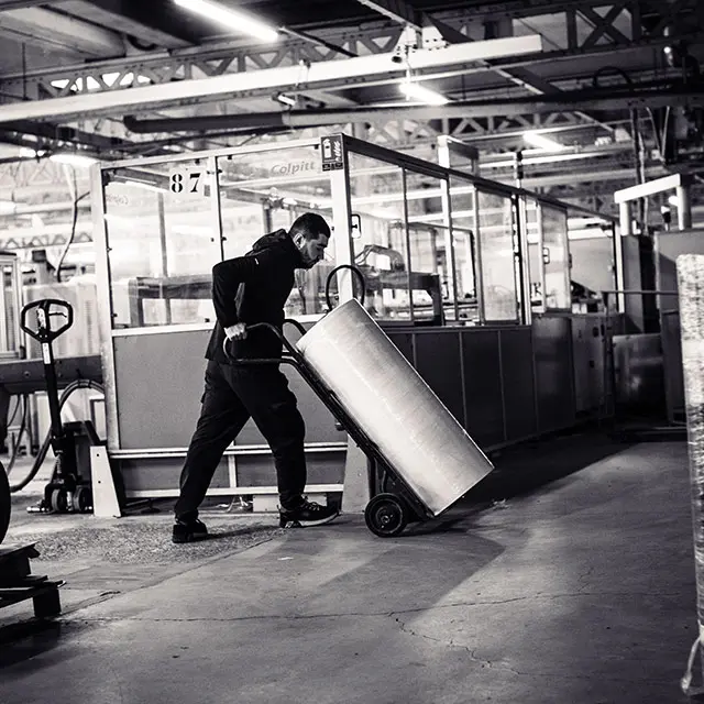Employé poussant un transpalette incliné chargé d'un grand cylindre métallique dans un atelier industriel lumineux.