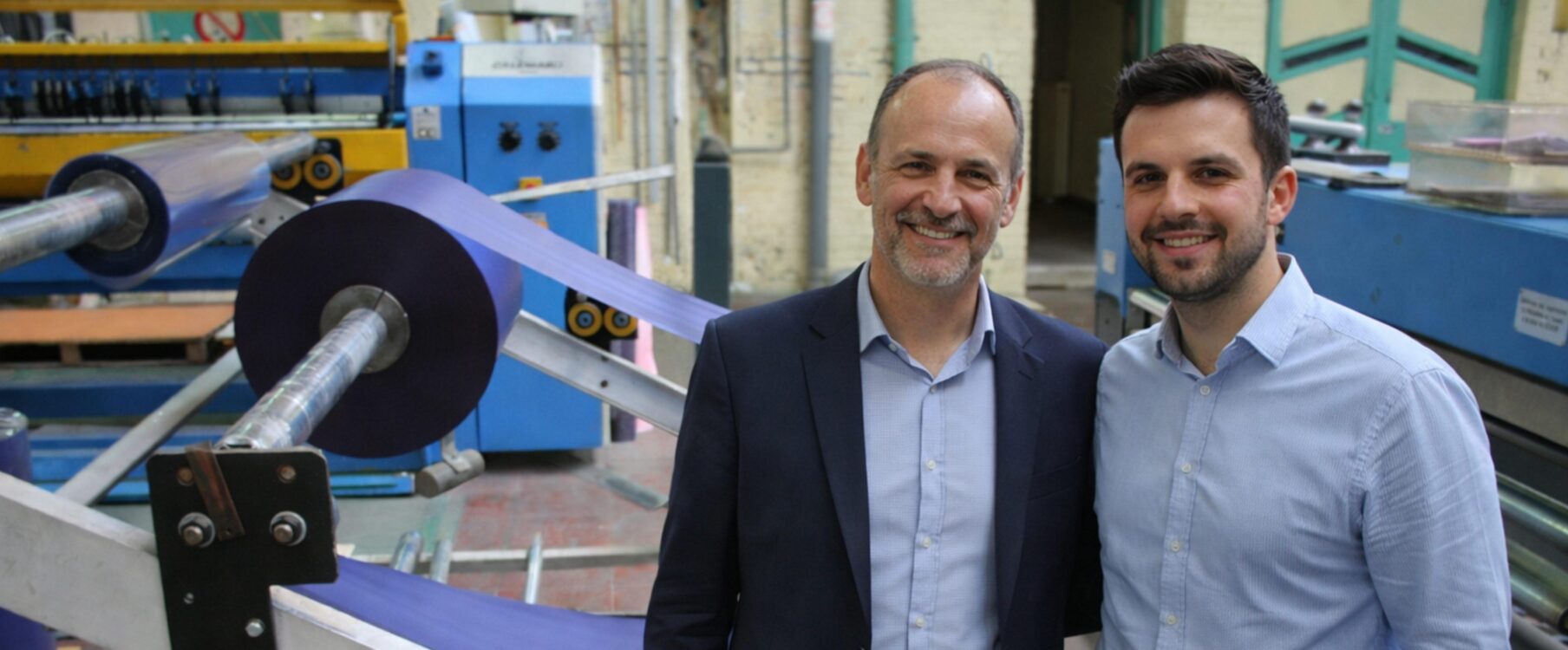 Deux hommes souriants en chemise et veste debout devant une machine industrielle enroulant une grande bobine de film plastique violet.