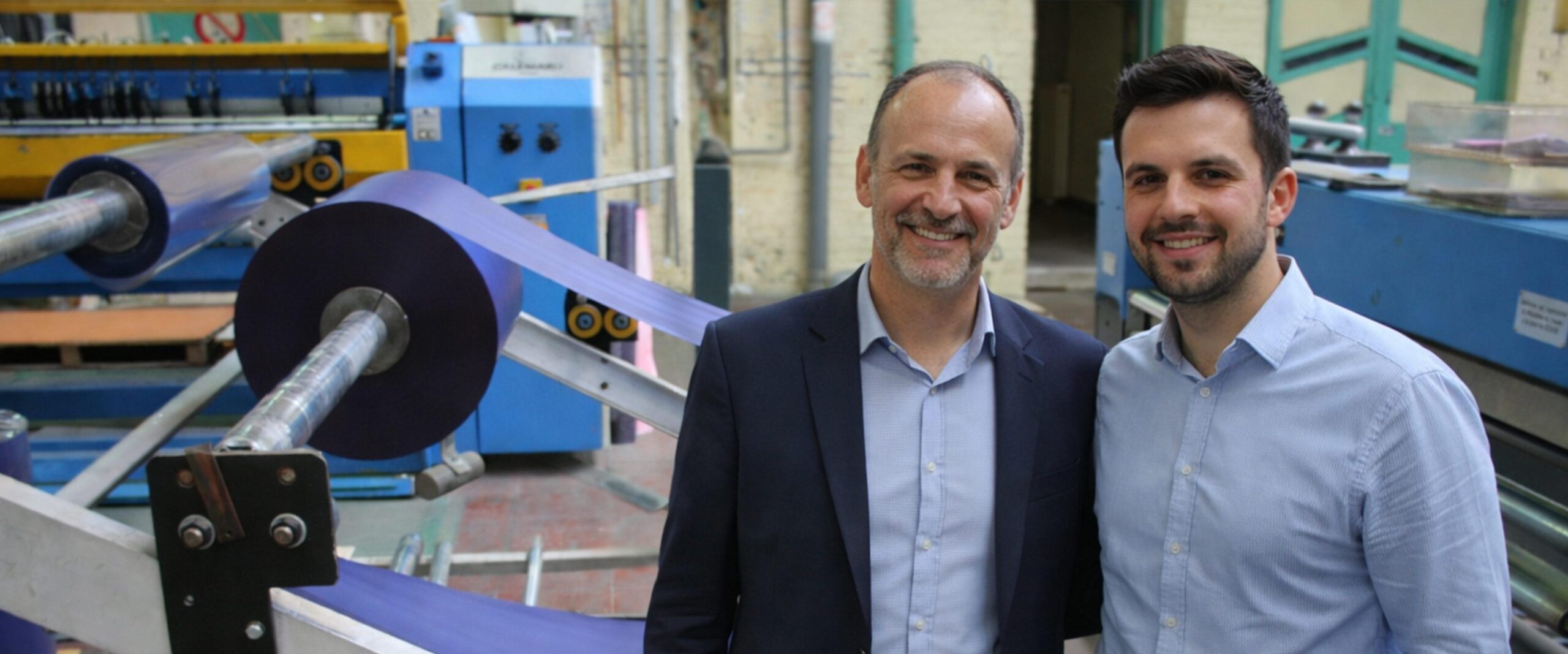 Deux hommes souriants en chemise et veste debout devant une machine industrielle enroulant une grande bobine de film plastique violet.